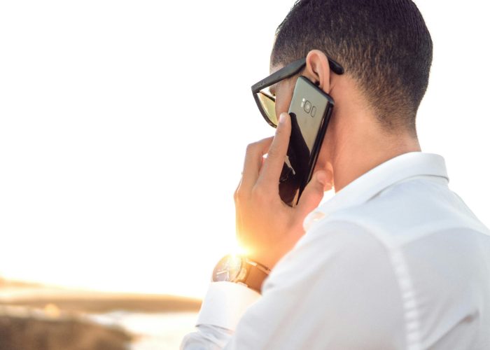 Man outdoors in Morocco talking on smartphone, wearing sunglasses and white shirt, backlit by sunset.